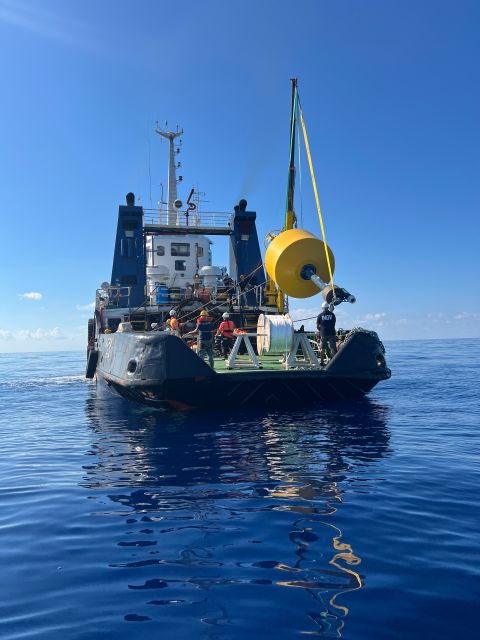 A dark vessel's stern, with crew members deploying a large yellow cylindrical buoy into the calm blue ocean using a crane under a clear sky.