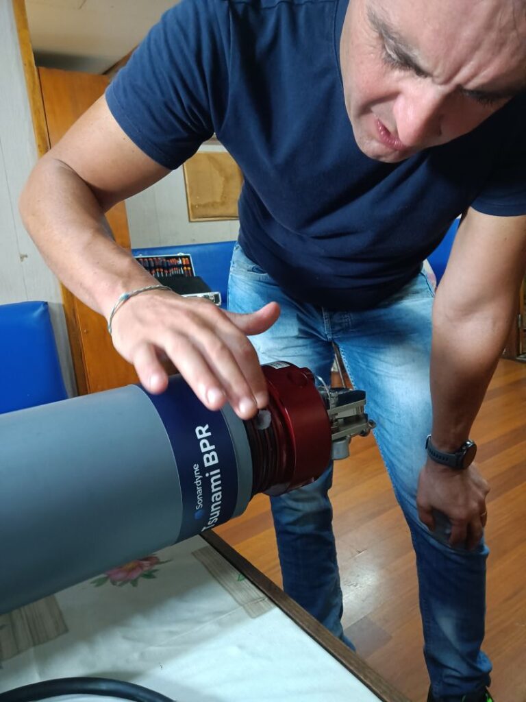 A man in a blue t-shirt and jeans inspects a gray and red Sonardyne Tsunami BPR cylindrical device on a table.