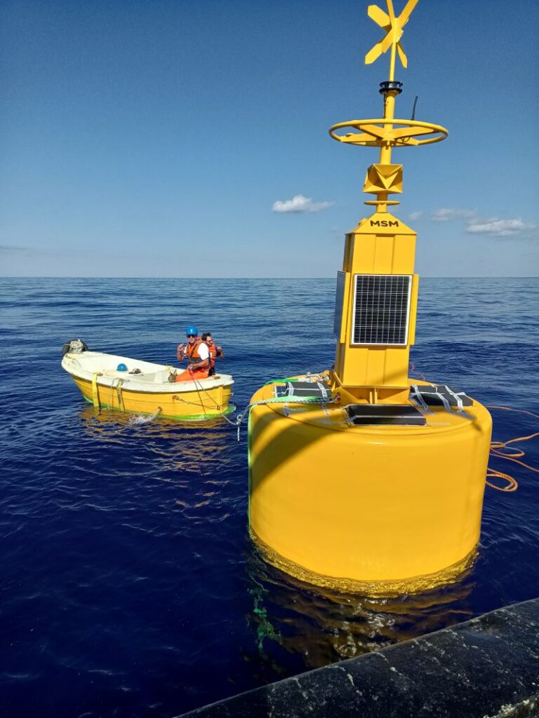 A large yellow oceanographic buoy with solar panels and a wind turbine floats next to a small boat with two people in life vests on a calm blue ocean.