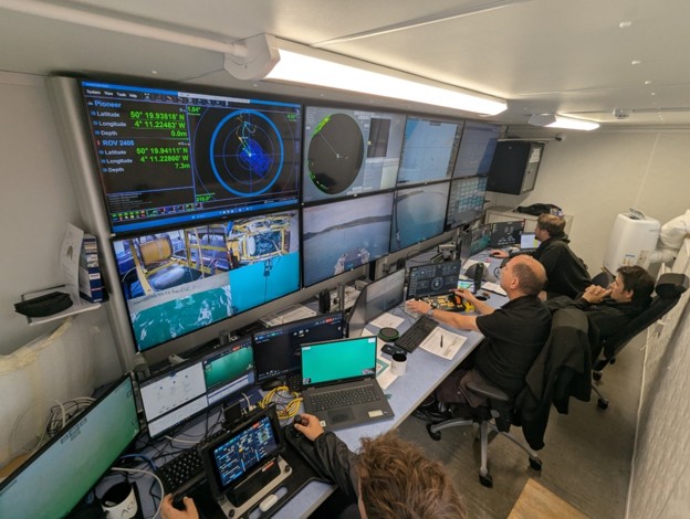 Four people in a brightly lit control room, surrounded by multiple computer screens displaying navigation, sonar, and live underwater footage of an ROV.