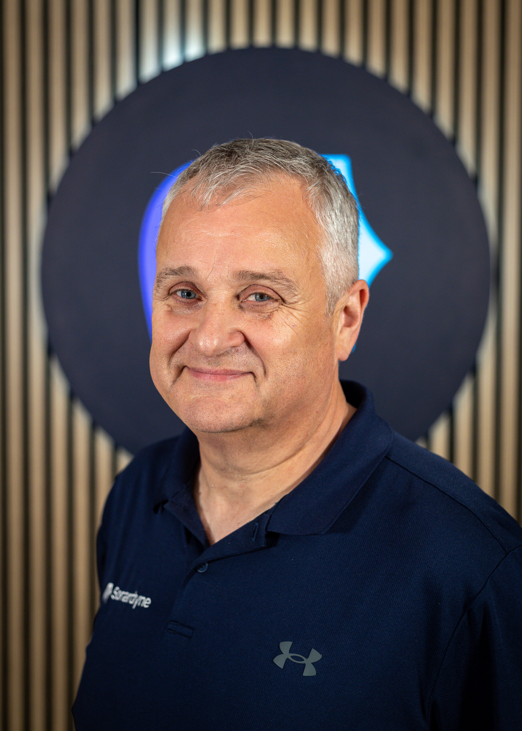 Headshot of a smiling man with grey hair in a dark blue polo shirt with "Sorardyne" and Under Armour logos, against a dark blue and wood-paneled background.