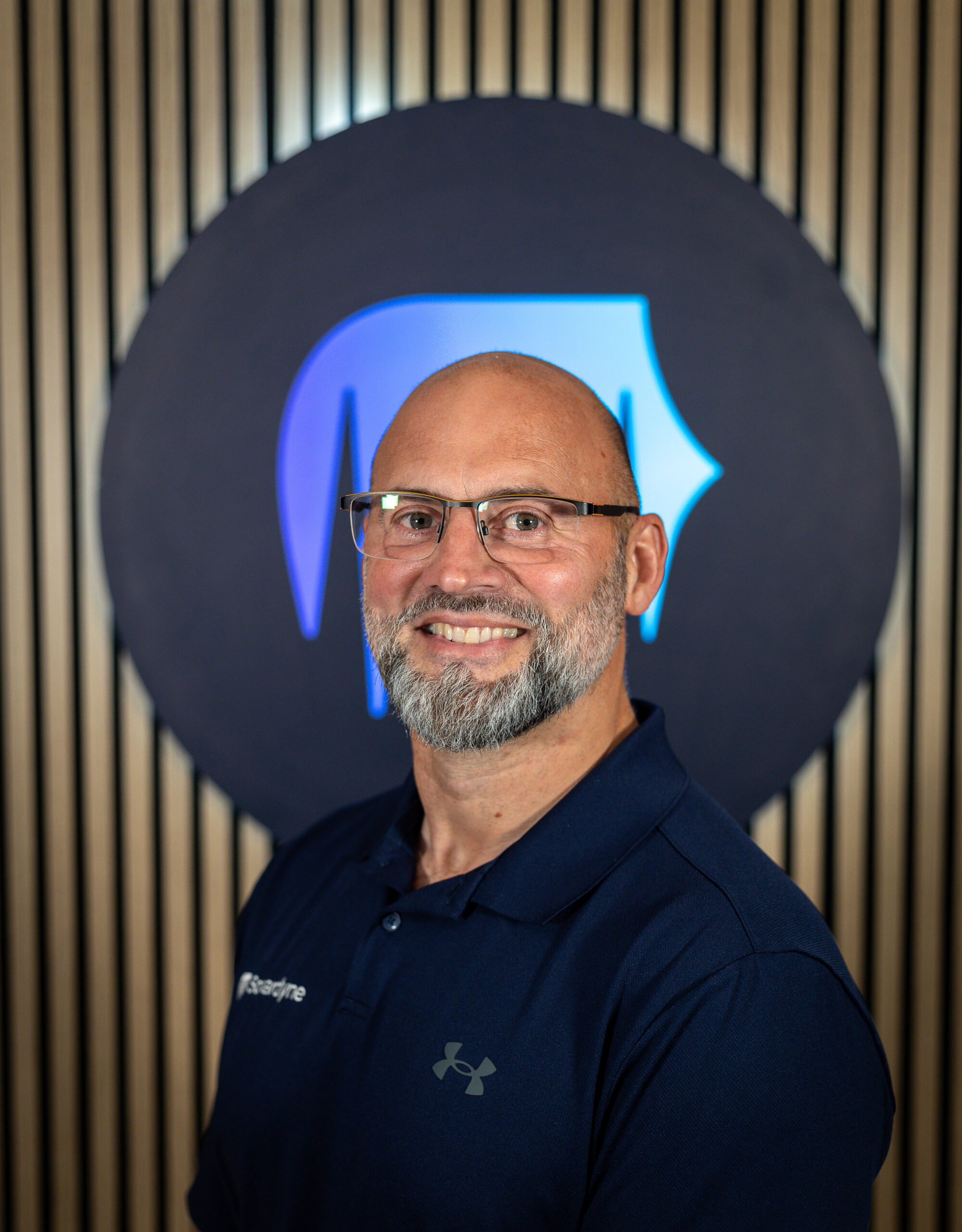 Headshot of a smiling bald man with a gray beard and glasses, wearing a dark blue polo shirt, against a background with a blue abstract logo and wooden slats.