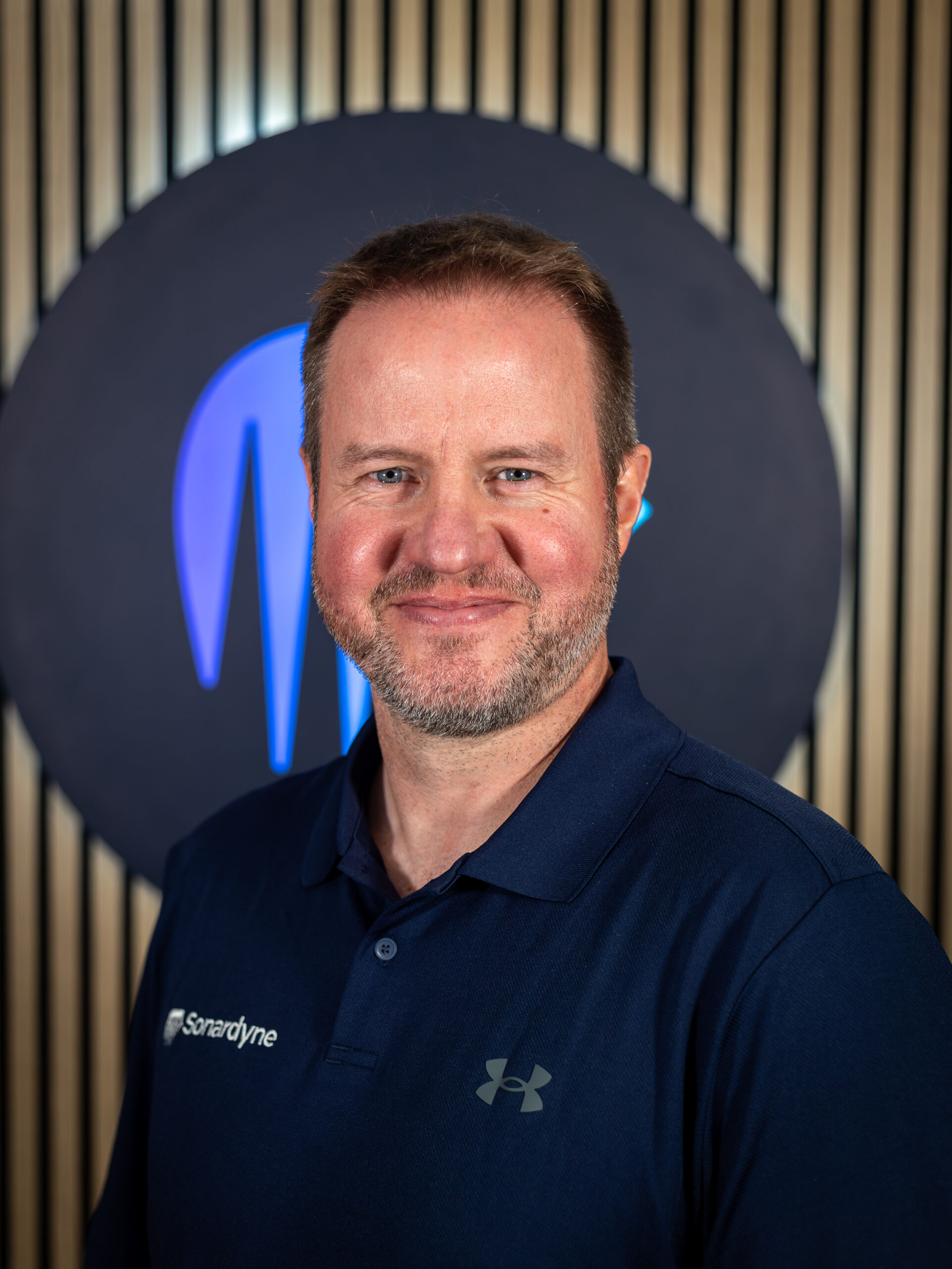 Headshot of a smiling man with blue eyes and a beard, wearing a dark blue Sonardyne polo shirt. A blue glowing logo is visible in the background.