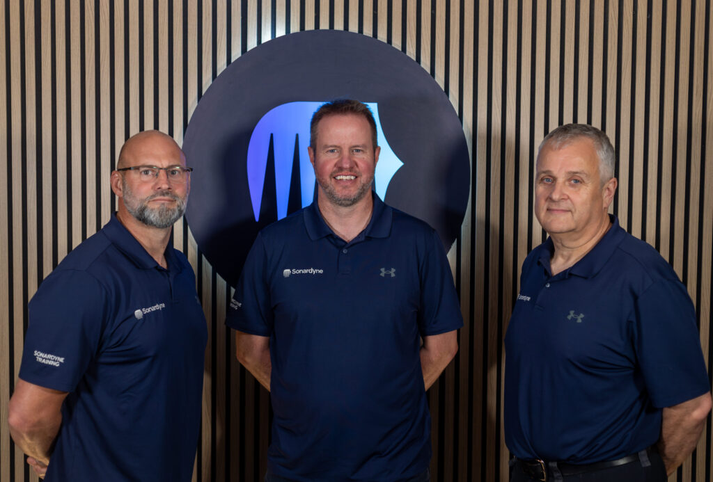 Three men in dark blue Sonardyne polo shirts stand in front of a wooden slatted wall with a large circular logo behind them.