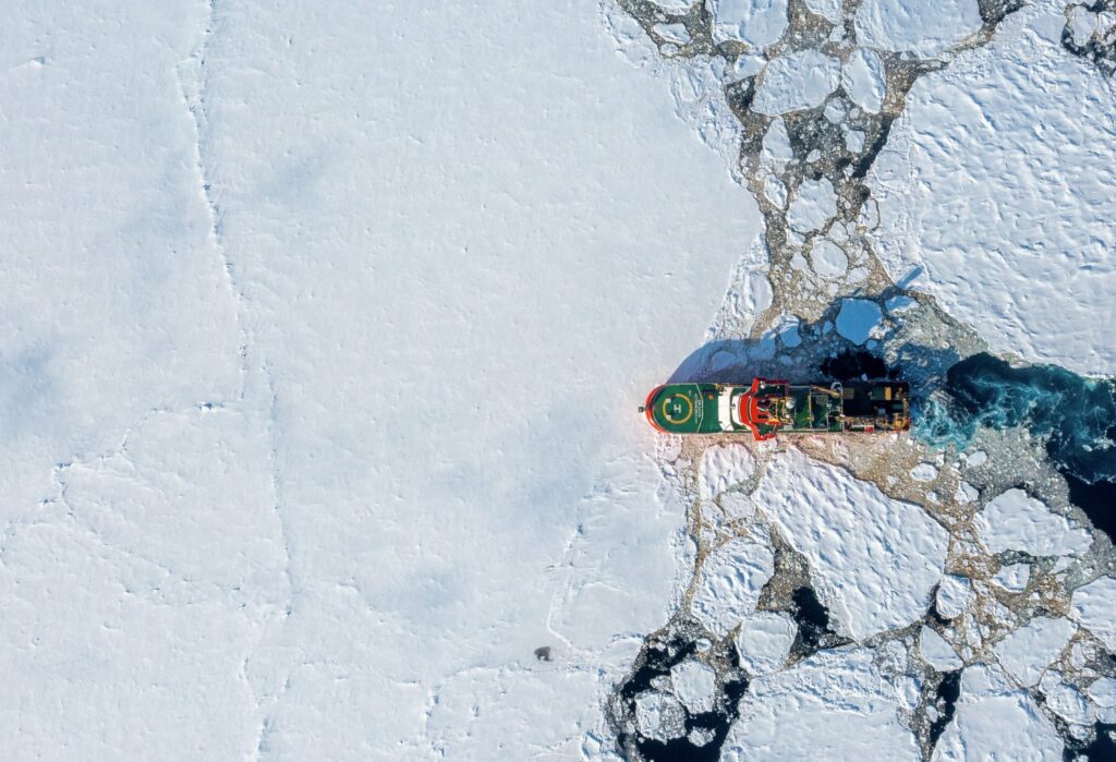 Aerial view of a red and green icebreaker ship breaking through vast white sea ice, with a small polar bear visible on the ice to the left.